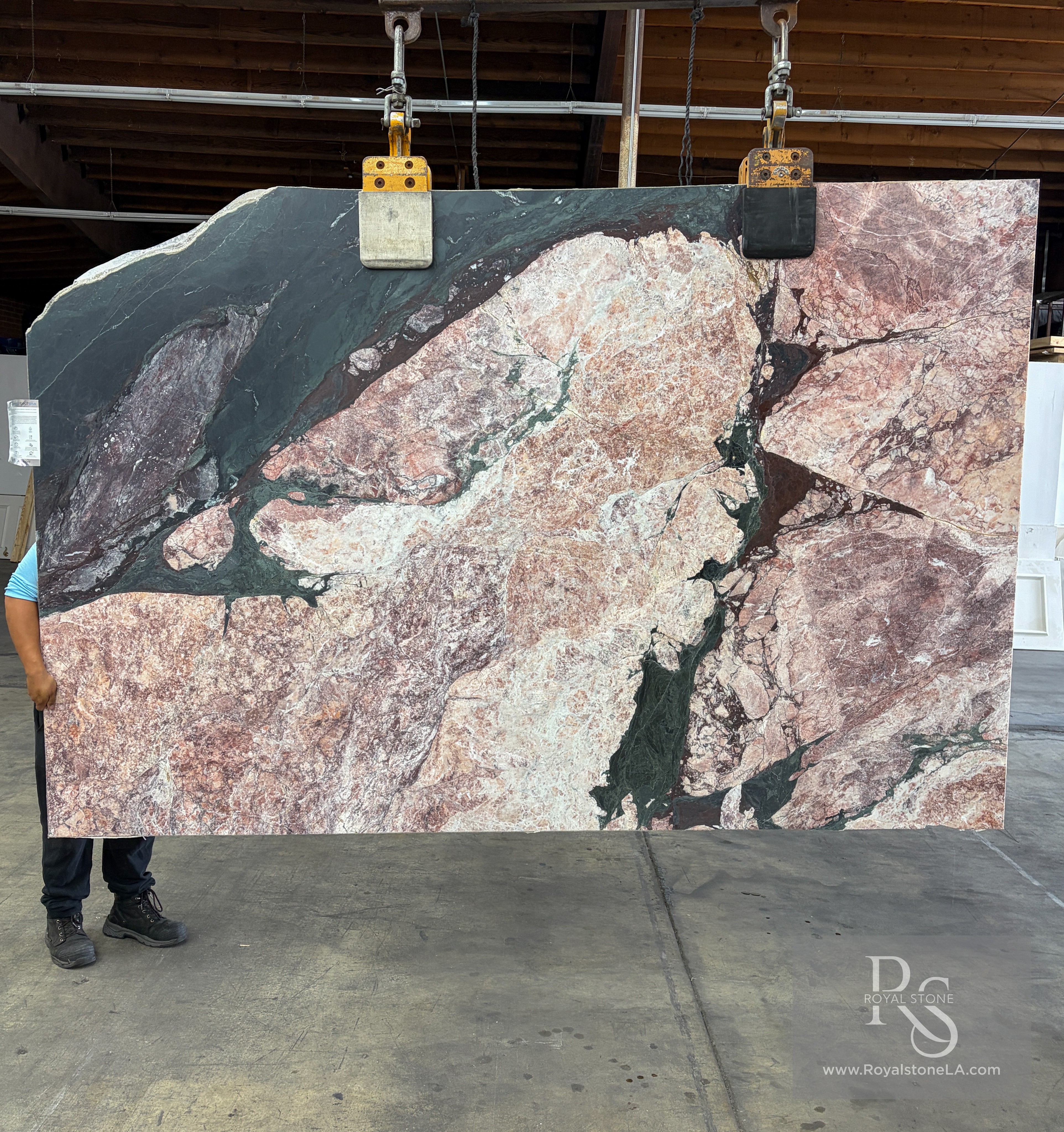 Antolini Opera D'Arte Select marble slab, 2cm honed, 74 by 119 inches, with rose-and-cream field, oxblood veining, and a dramatic forest-green serpentine corner, hanging from a crane at the Royal Stone Los Angeles yard