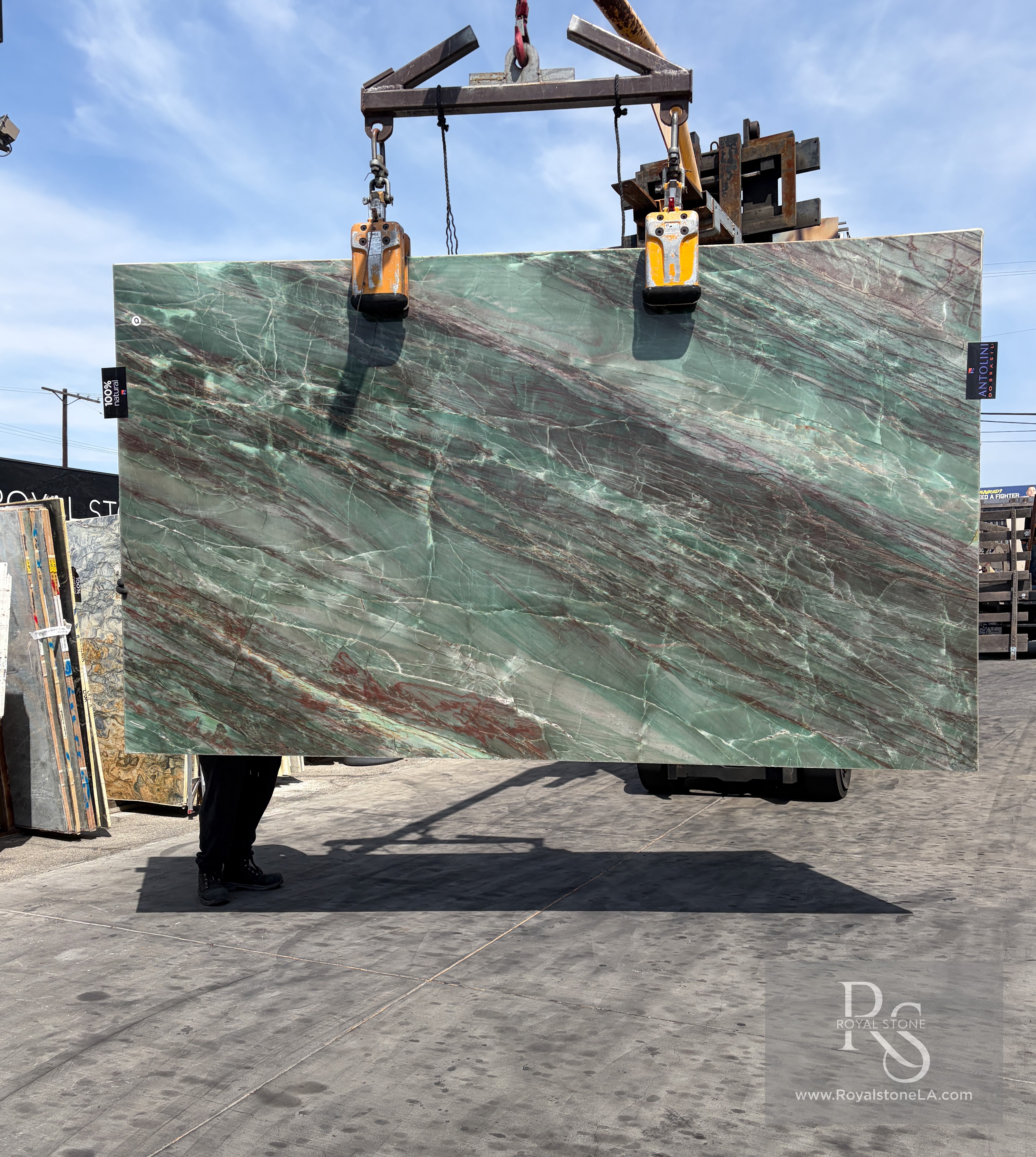 Antolini Thunderflash green quartzite slab, 2cm polished, 76 by 129 inches, with sweeping diagonal red and copper veining across a pale seafoam-green field, hanging from a crane at the Royal Stone Los Angeles yard